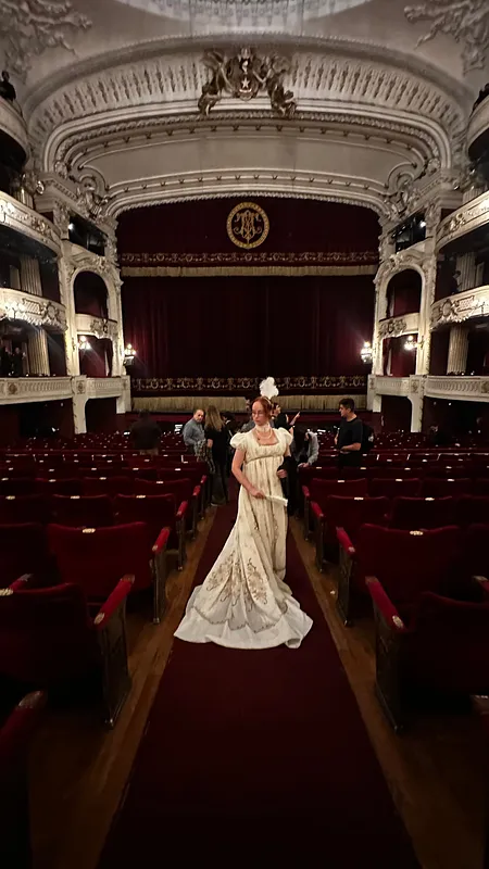 Vista en tres cuartos de una persona con vestido blanco de falda amplia y cola, posando entre las butacas de un teatro.