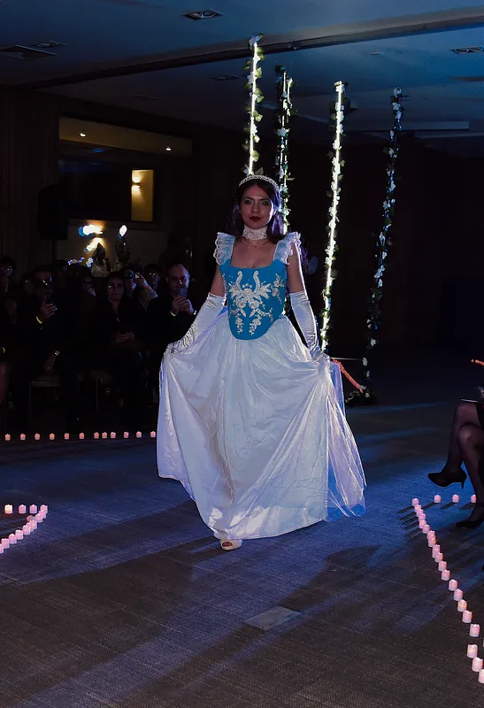 Modelo caminando en pasarela oscura con vestido celeste y blanco, guantes largos, gargantilla, tiara y falda tomada a ambos lados para mostrar el volumen.