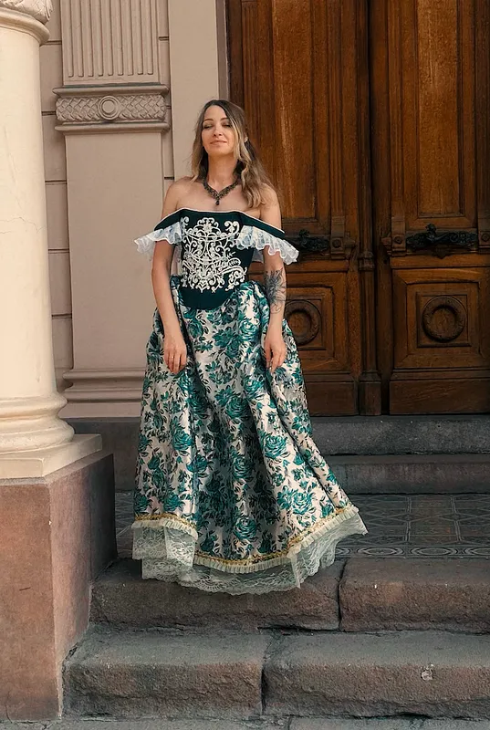 Vista frontal de una mujer con vestido largo de época, cuerpo oscuro bordado en claro, escote hombros descubiertos con encaje blanco y falda floral amplia en tonos verde y marfil.