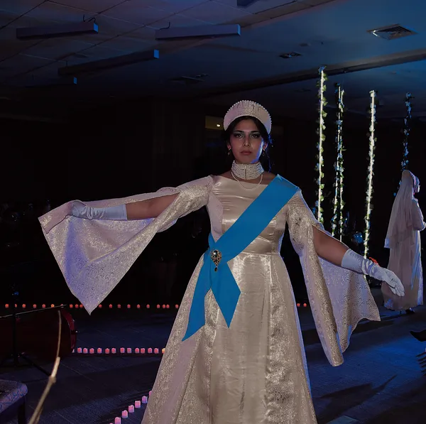Modelo con los brazos extendidos mostrando las mangas abiertas y brocadas del vestido marfil, con banda celeste, guantes y corona bajo luz azul.