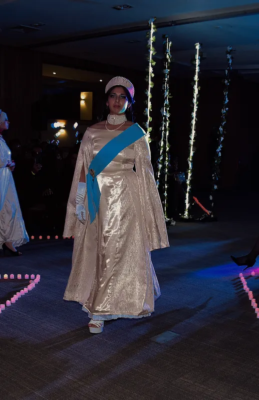 Vista completa en pasarela oscura del vestido Reina marfil, combinado con corona, gargantilla, guantes blancos y banda celeste cruzada.