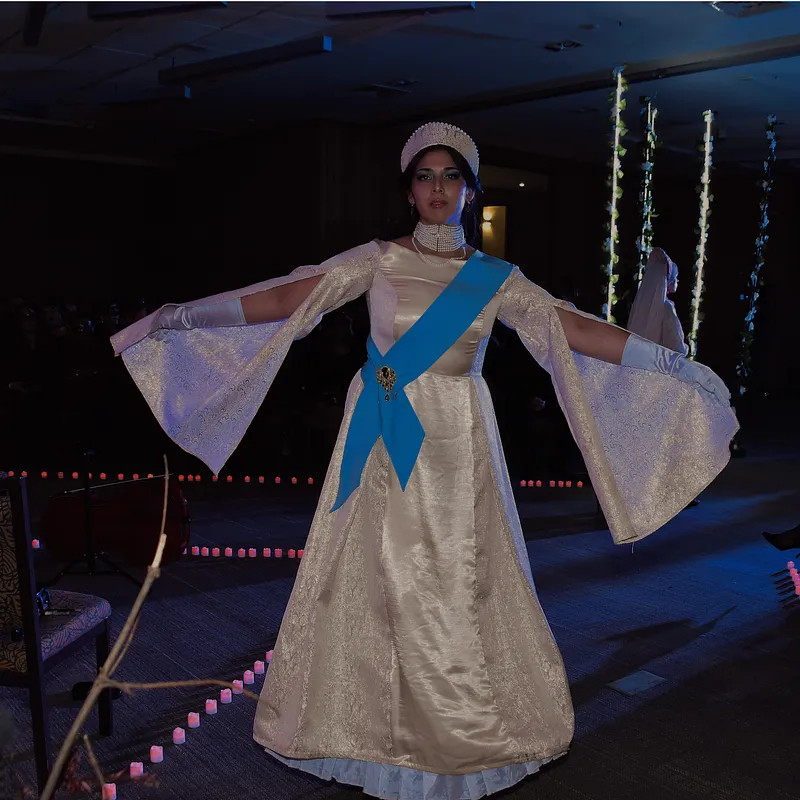 Vista frontal amplia del vestido Reina en pasarela, destacando la falda larga, el brillo satinado, las mangas acampanadas y la banda celeste.