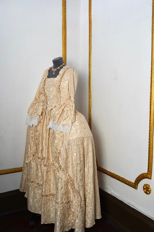 Vista frontal en tres cuartos del vestido, mostrando el volumen lateral de la falda, la sobrefalda con vuelos diagonales y las mangas acampanadas con puños de encaje.