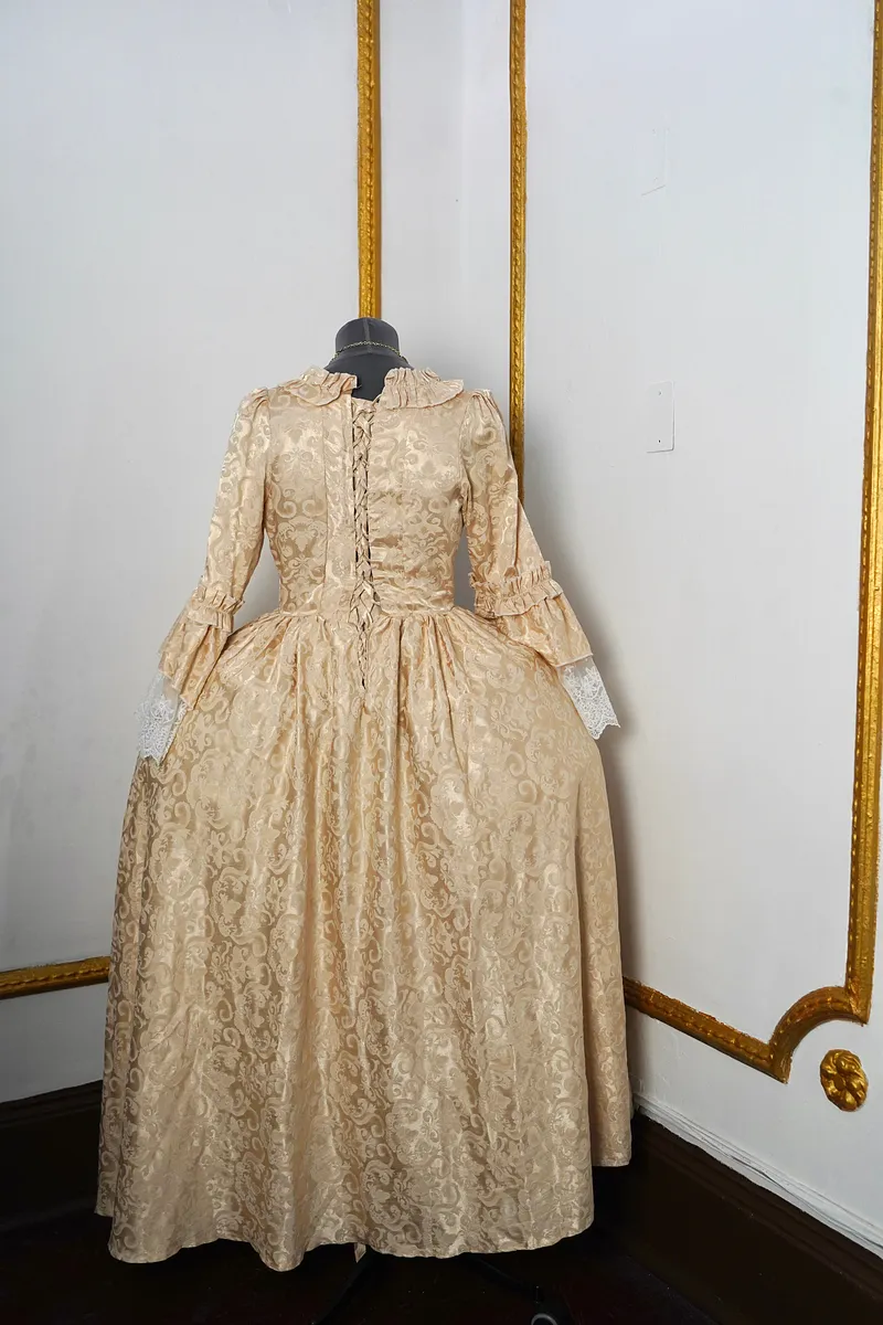 Vista posterior del vestido en maniquí, con espalda ajustada, cierre acordonado, cintura marcada, falda amplia de brocado y terminaciones de encaje visibles en las mangas.