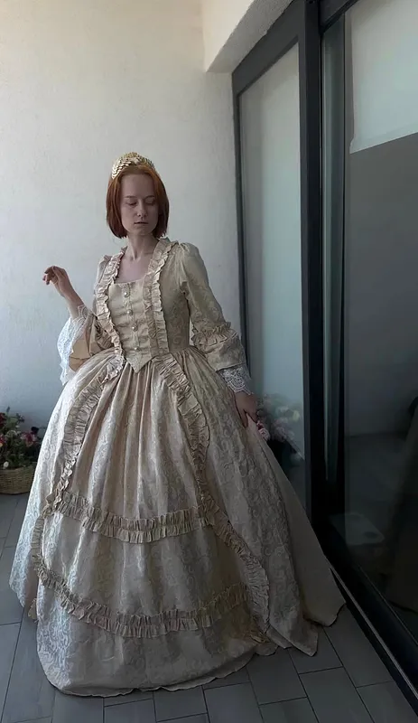 Vestido Charlotta usado por una modelo, estilizado con tocado dorado; se aprecia la caída de la falda, el escote cuadrado, los botones frontales, vuelos y encaje en las mangas.