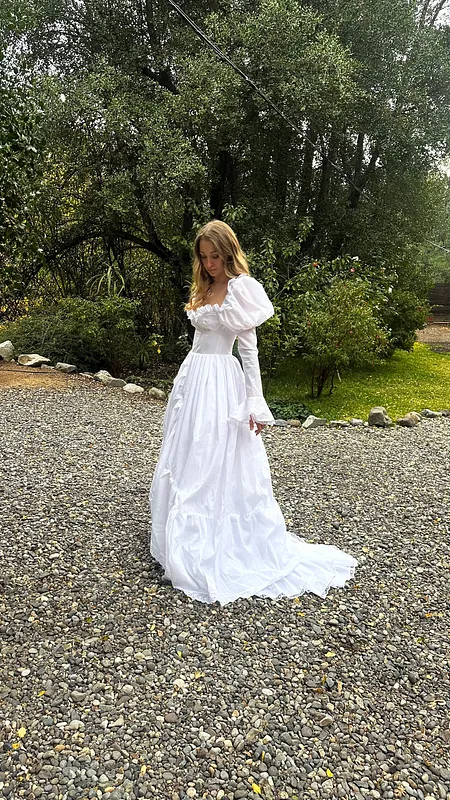Vista completa a distancia del vestido blanco, con cuerpo ajustado, falda de gran volumen y cola arrastrándose sobre la grava.