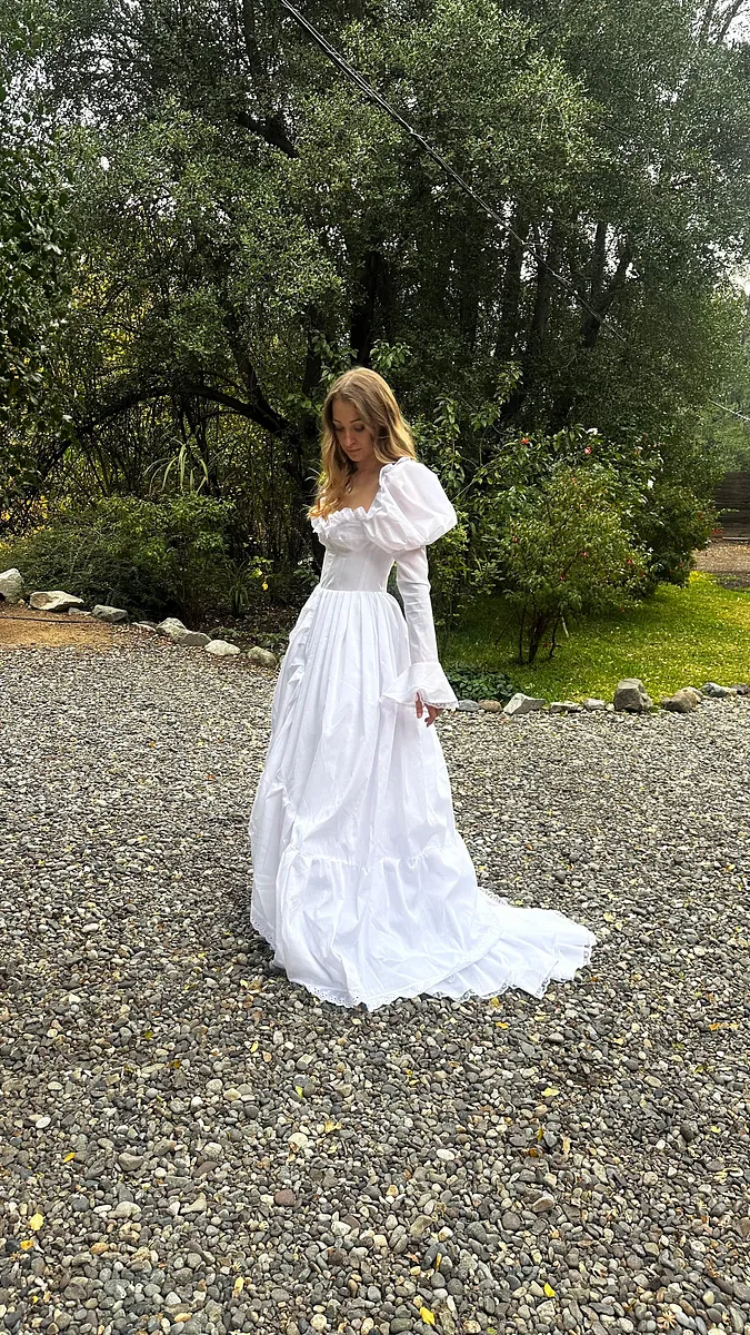 Vista completa a distancia del vestido blanco, con cuerpo ajustado, falda de gran volumen y cola arrastrándose sobre la grava.