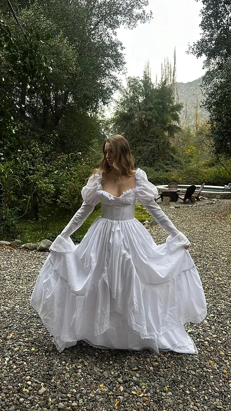 Vista frontal al aire libre de un vestido blanco amplio, con escote cuadrado, mangas largas y falda recogida por la modelo.
