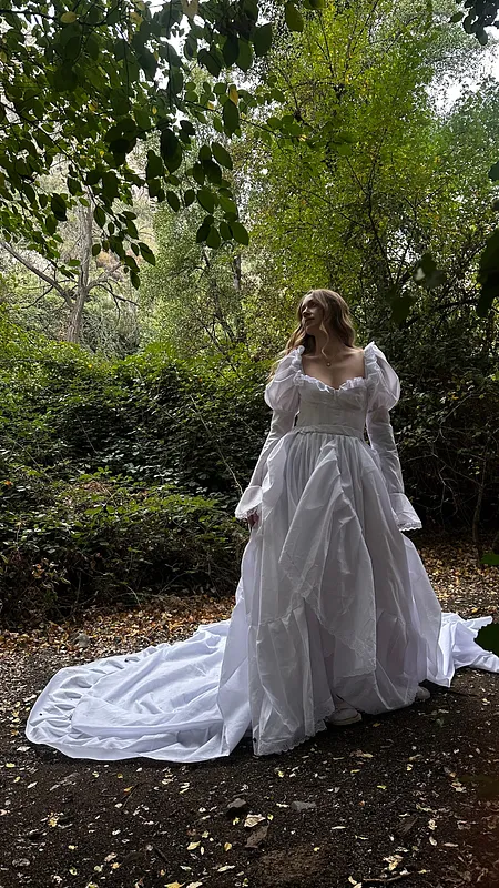 Pose de tres cuartos bajo los árboles, con vestido blanco de mangas largas, falda voluminosa y amplia cola desplegada.