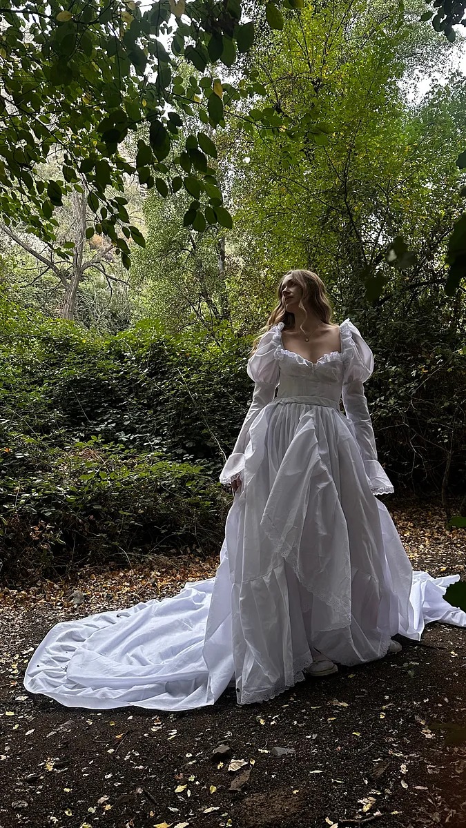 Pose de tres cuartos bajo los árboles, con vestido blanco de mangas largas, falda voluminosa y amplia cola desplegada.