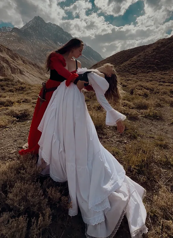 Escena en la montaña con una mujer de falda blanca y corpiño oscuro sostenida por otra persona vestida de rojo.