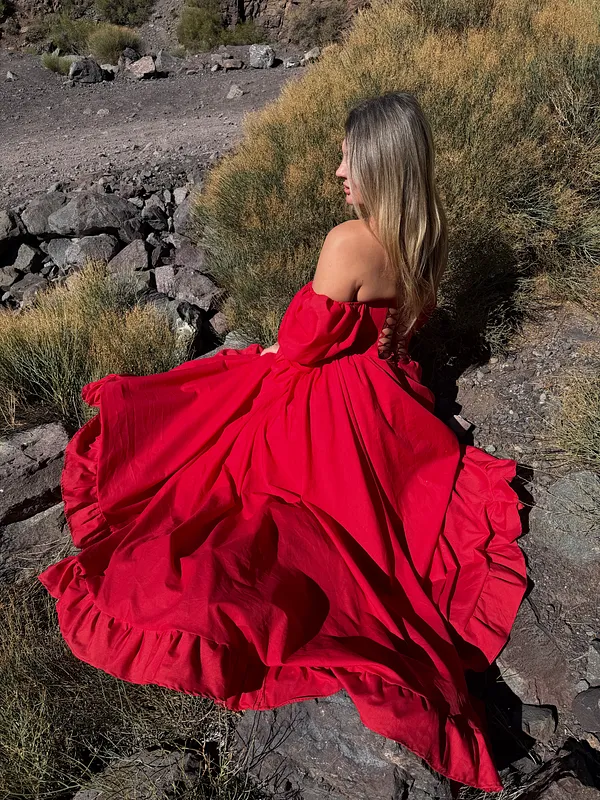 Vista posterior de una mujer sentada sobre rocas con vestido rojo de hombros descubiertos y falda extendida.