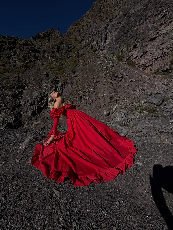 Mujer con vestido rojo de gran vuelo levanta la falda mientras posa de perfil en un paisaje montañoso y rocoso.