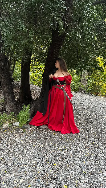 Mujer de pie bajo un árbol luce vestido rojo de escote amplio, mangas caídas y larga falda sobre un sendero.