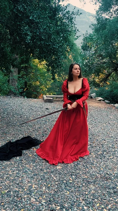 Mujer con vestido rojo posa en un camino arbolado sosteniendo una espada, con una capa negra extendida detrás.