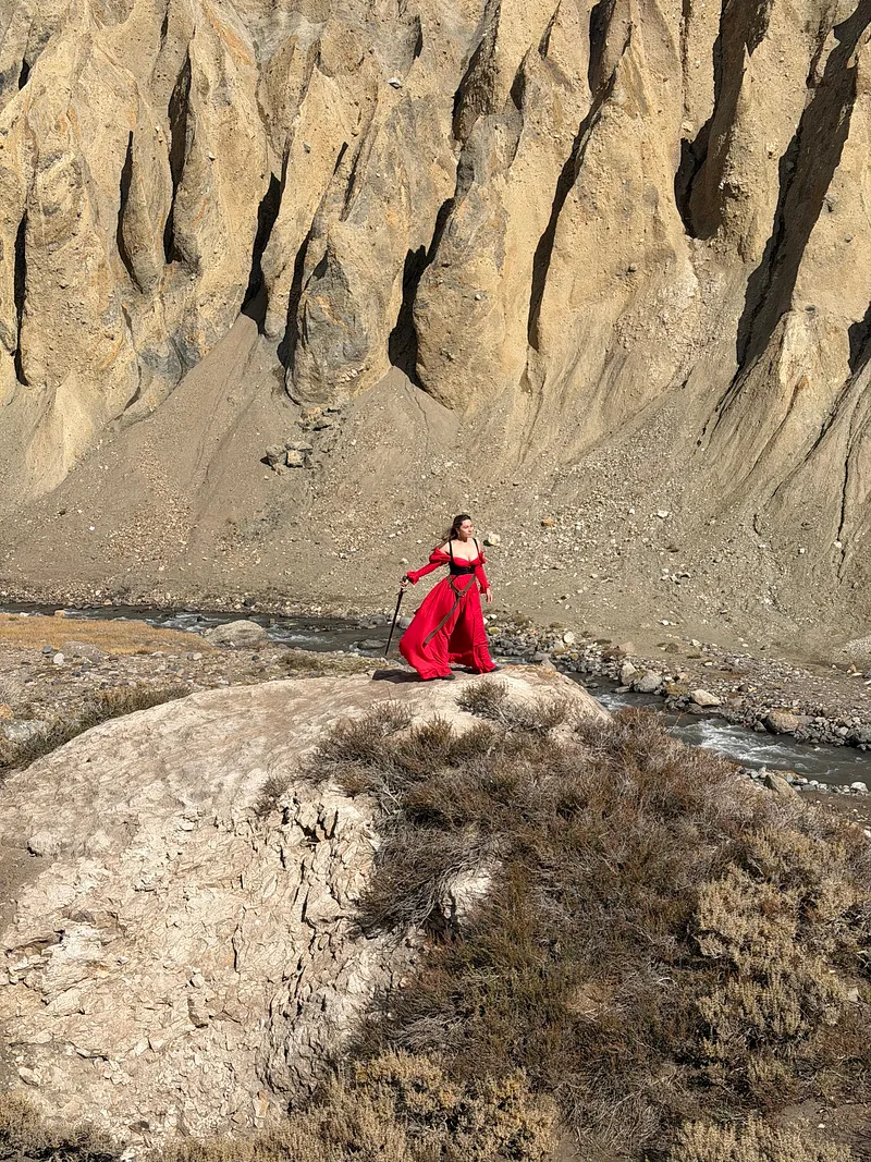 Vista panorámica de una mujer con vestido rojo junto a un río, rodeada por grandes paredes de roca.