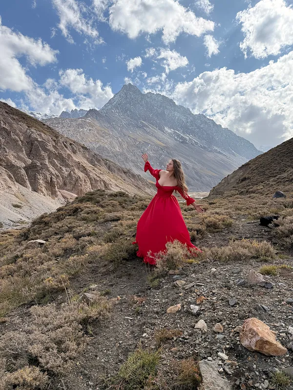Mujer vista de espaldas camina por un valle rocoso con vestido rojo, bajo un cielo azul con nubes.