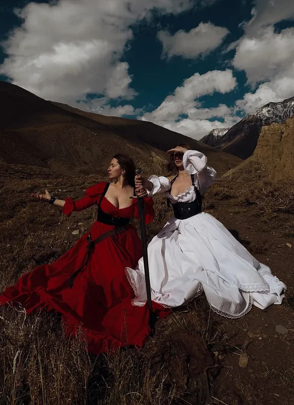 Dos mujeres con vestidos rojo y blanco avanzan entre montañas, con las faldas desplegadas por el terreno.