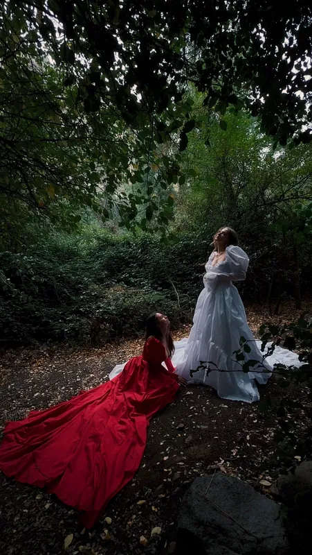 Dos mujeres con vestidos rojo y blanco posan juntas en un bosque; la larga cola roja se extiende sobre el suelo.
