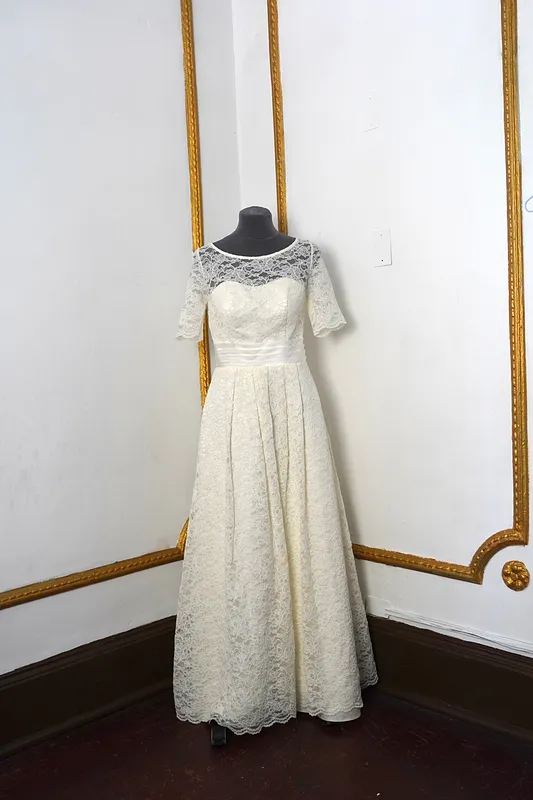 Vista frontal del vestido en maniquí: escote ilusión de encaje, mangas cortas festoneadas, cintura satinada y falda larga amplia.