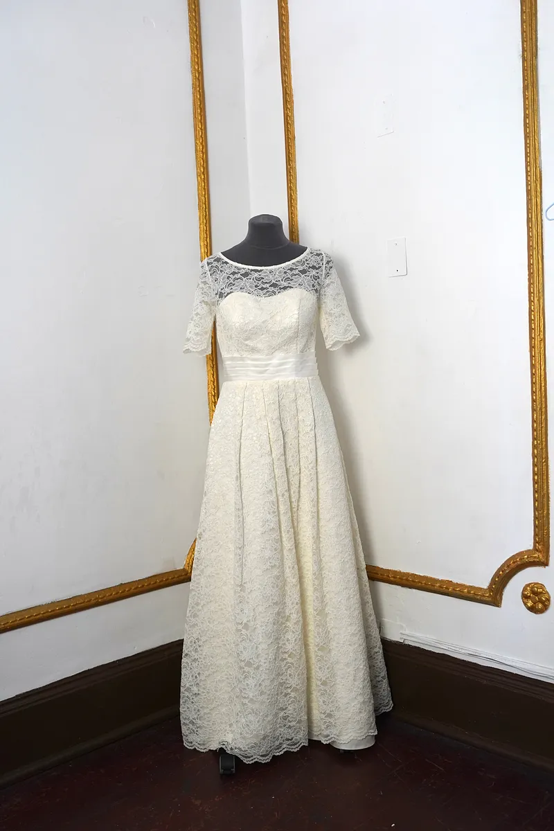 Vista frontal del vestido en maniquí: escote ilusión de encaje, mangas cortas festoneadas, cintura satinada y falda larga amplia.