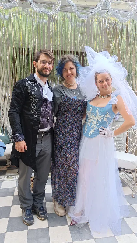 Tres personas posan en un patio; la mujer de la derecha viste el corsé celeste bordado con falda de tul blanco, guantes de encaje, collar y gran tocado de tul.