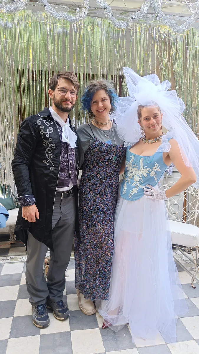 Tres personas posan en un patio; la mujer de la derecha viste el corsé celeste bordado con falda de tul blanco, guantes de encaje, collar y gran tocado de tul.