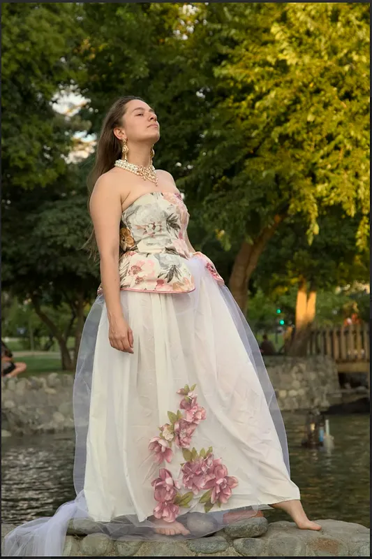 Vista exterior del vestido Peoni sobre piedras junto al agua, mostrando el cuerpo floral, la falda blanca amplia, tul transparente y aplicaciones florales rosadas.