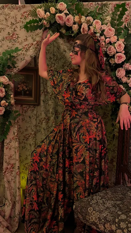 Mujer posando junto a un muro cubierto de rosas, con vestido largo oscuro de estampado floral rojo y mangas abullonadas.