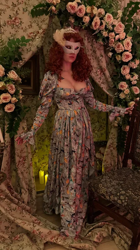 Modelo con peluca rojiza y antifaz blanco luce el vestido floral en un decorado romántico con cortinas estampadas, rosas y luz cálida.