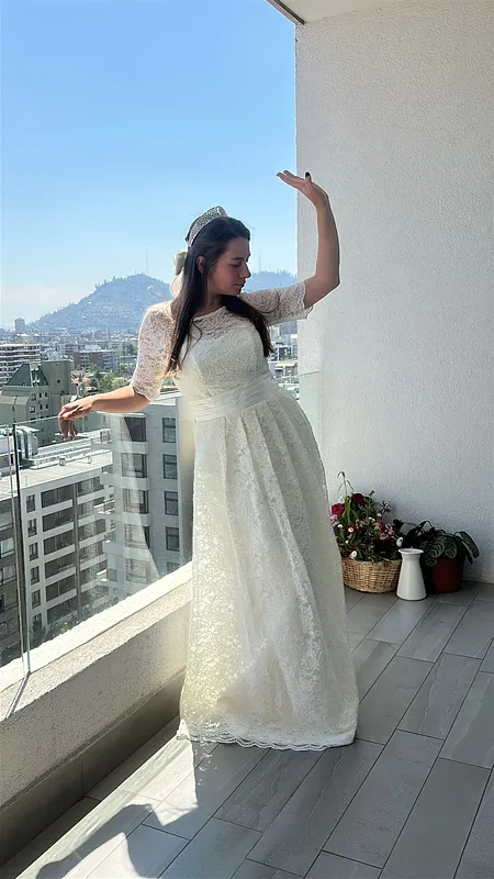 Mujer usando vestido largo de novia en encaje marfil, con manga corta transparente, cinturón satinado y tiara, posando en un balcón.