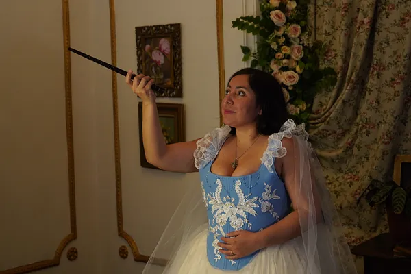 Mujer con vestido de corsé celeste bordado, falda blanca de tul y velo, sosteniendo una varita en un interior decorado con flores, cuadros y molduras doradas.