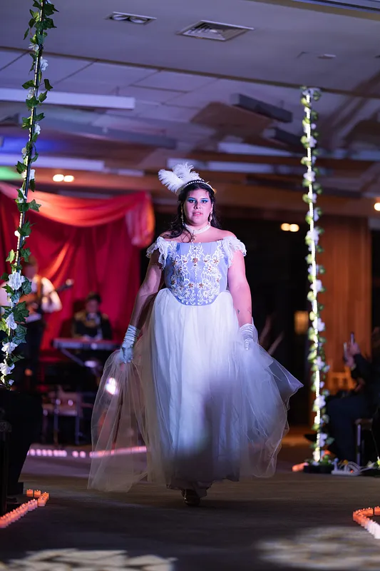 Modelo caminando en pasarela con vestido lila y blanco de falda amplia de tul, corpiño bordado, guantes claros y tocado de plumas bajo luces de escenario.