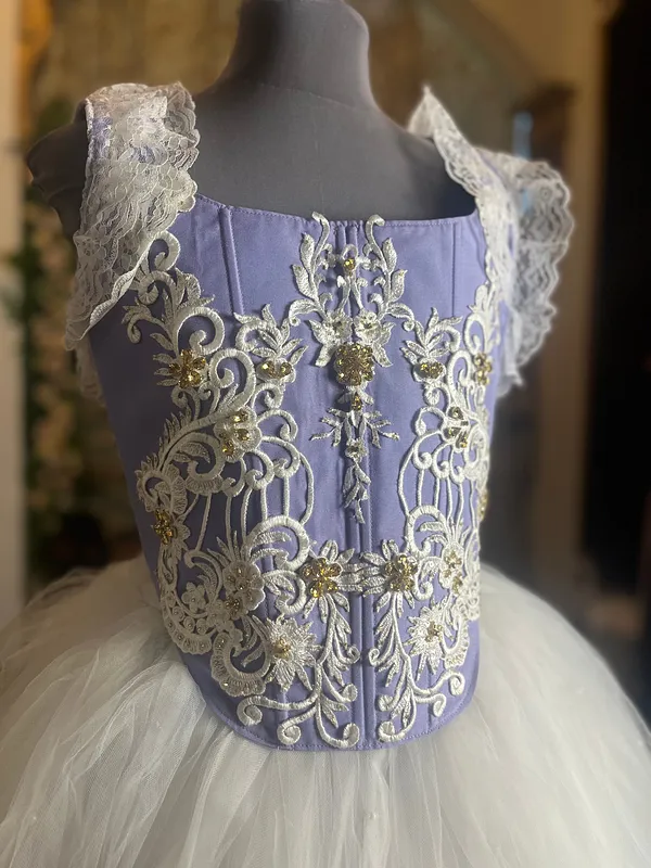 Acercamiento del corpiño lila con costuras verticales, bordados ornamentales marfil, pedrería dorada, escote cuadrado y tirantes anchos de encaje.