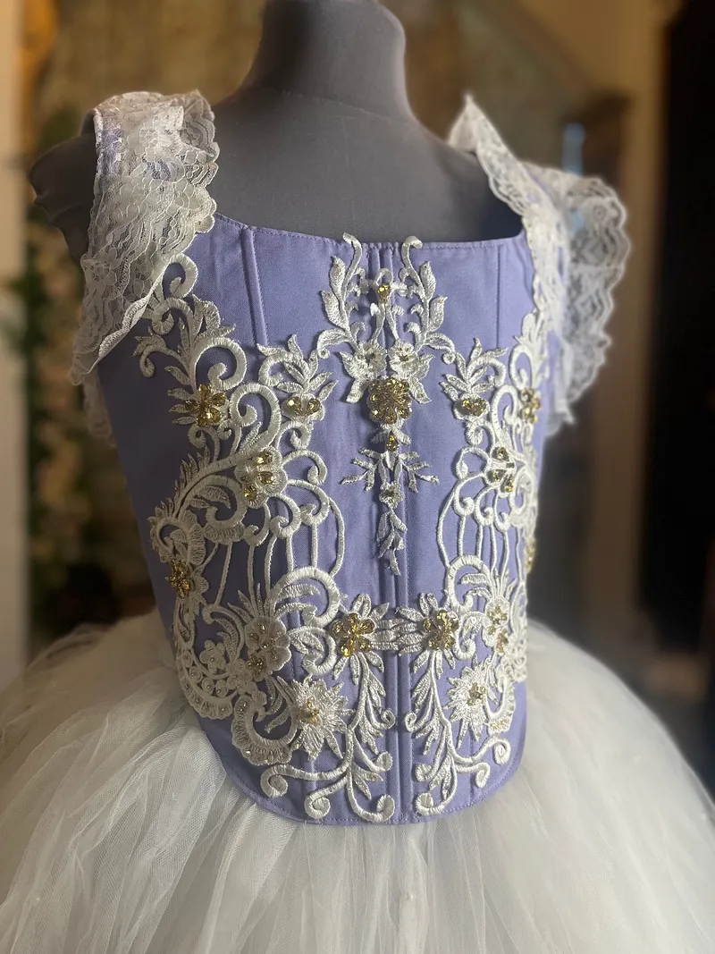 Acercamiento del corpiño lila con costuras verticales, bordados ornamentales marfil, pedrería dorada, escote cuadrado y tirantes anchos de encaje.