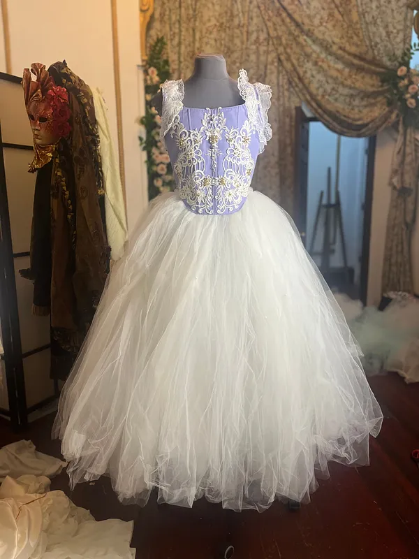 Vista frontal del vestido sobre maniquí, con corpiño lila bordado en marfil y dorado, tirantes de encaje blanco y falda voluminosa de tul.