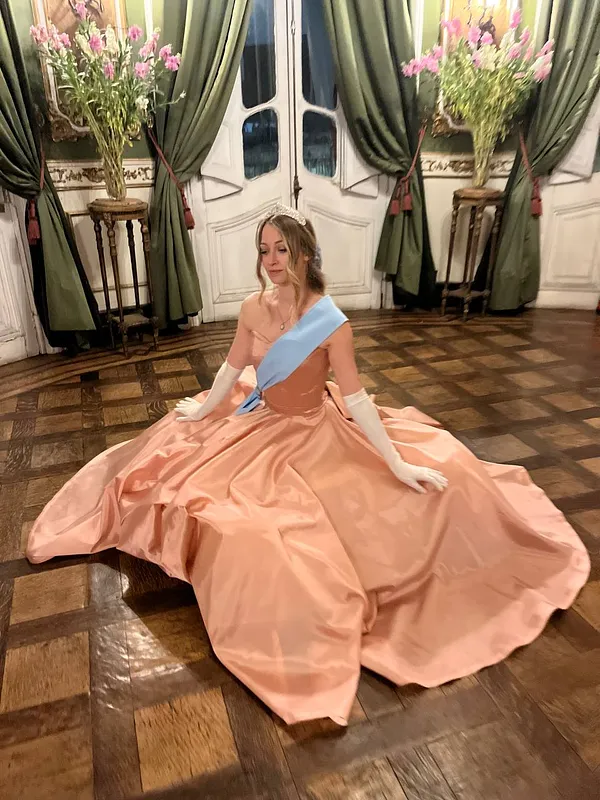 Mujer sentada en un salón con vestido de baile color durazno, falda satinada muy amplia, banda celeste y guantes blancos.
