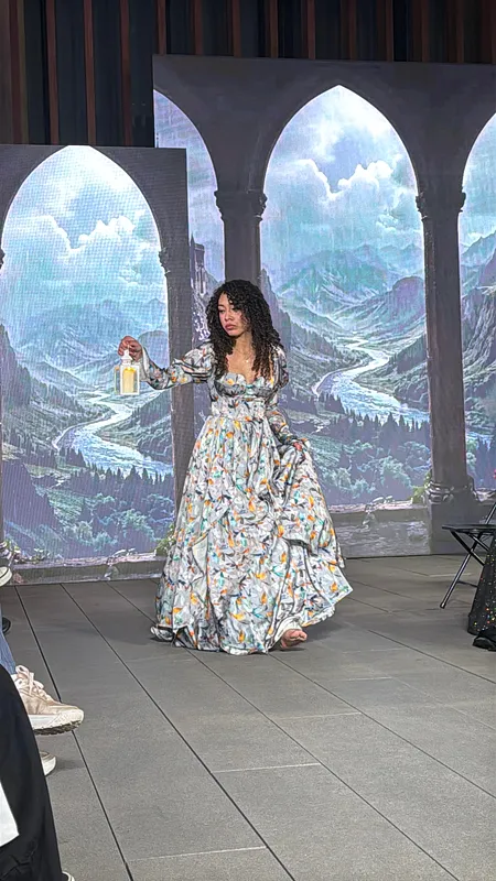 Modelo posa en escenario con vestido floral amplio, escote cuadrado y mangas largas, sosteniendo una linterna frente a un telón de arcos y paisaje de montaña.