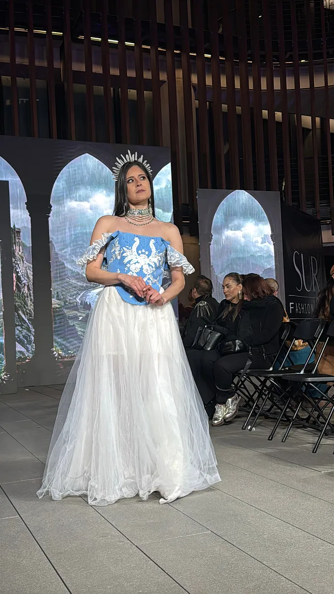Modelo de frente en pasarela con corsé celeste bordado, vuelos de encaje en los brazos, falda larga de tul blanco y tocado plateado tipo halo.