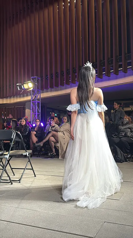 Vista posterior del vestido en pasarela, con corsé celeste sin tirantes visibles, vuelos de encaje en los brazos y falda blanca de tul con cola amplia.