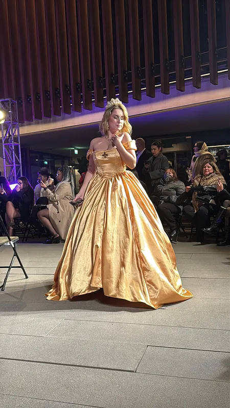 Mujer camina de frente por una pasarela con vestido dorado brillante, mangas caídas y falda amplia hasta el suelo.