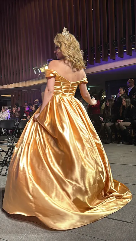 Vista posterior del vestido dorado en pasarela, con hombros descubiertos, falda recogida y una corta cola brillante.