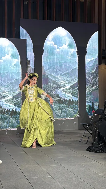 Bailarina en escenario usando el vestido verde con tocado, posando frente a paneles iluminados con arcos y paisaje; la falda larga cae en pliegues amplios.