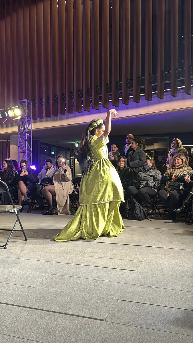 Vista lateral en pasarela de la bailarina usando el vestido verde; se aprecia la falda larga con arrastre, la cintura marcada y el tocado sobre el cabello recogido.