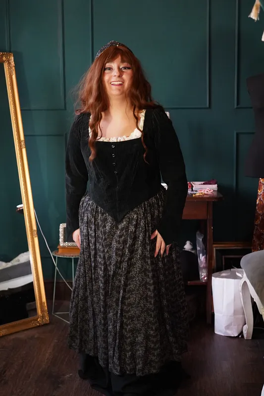 Vista frontal completa del vestido Katrin en uso, con cuerpo negro de mangas largas, escote con vuelos claros, falda floral oscura y capa inferior negra.