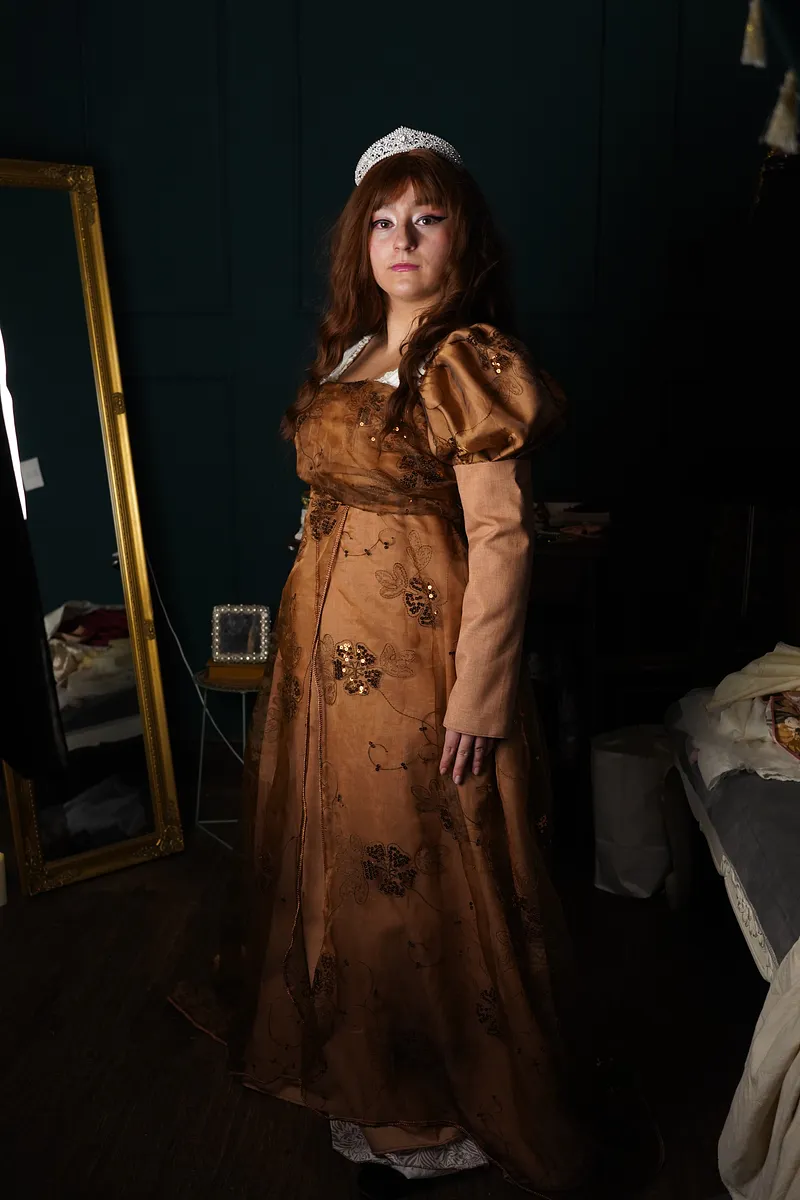 Vista de cuerpo completo del vestido Lilian en una modelo con tiara, mostrando la falda larga cobriza, mangas abullonadas y sobrefalda bordada en un interior oscuro.