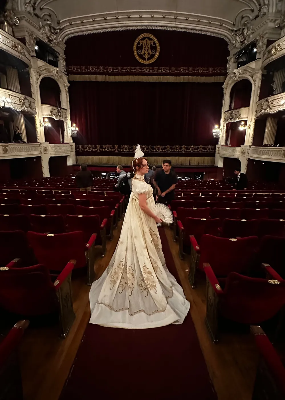 Vista posterior de una persona con vestido blanco largo y cola extendida, de pie en el pasillo central de un teatro.