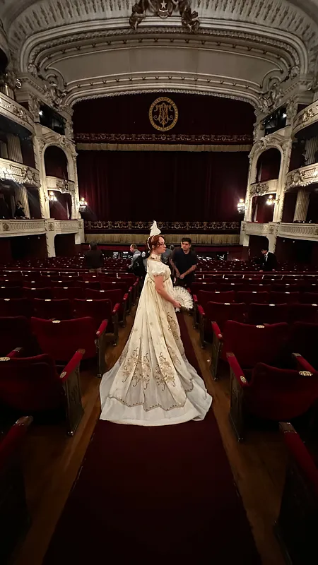 Vista posterior de una persona con vestido blanco largo y cola extendida, de pie en el pasillo central de un teatro.
