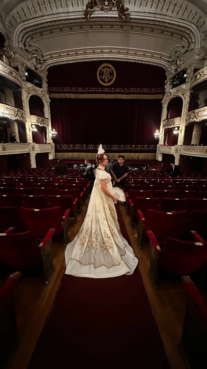 Vista posterior de una persona con vestido blanco largo y cola extendida, de pie en el pasillo central de un teatro.